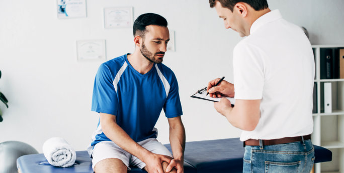 physiotherapist writing diagnosis while football player sitting on massage table in hospital